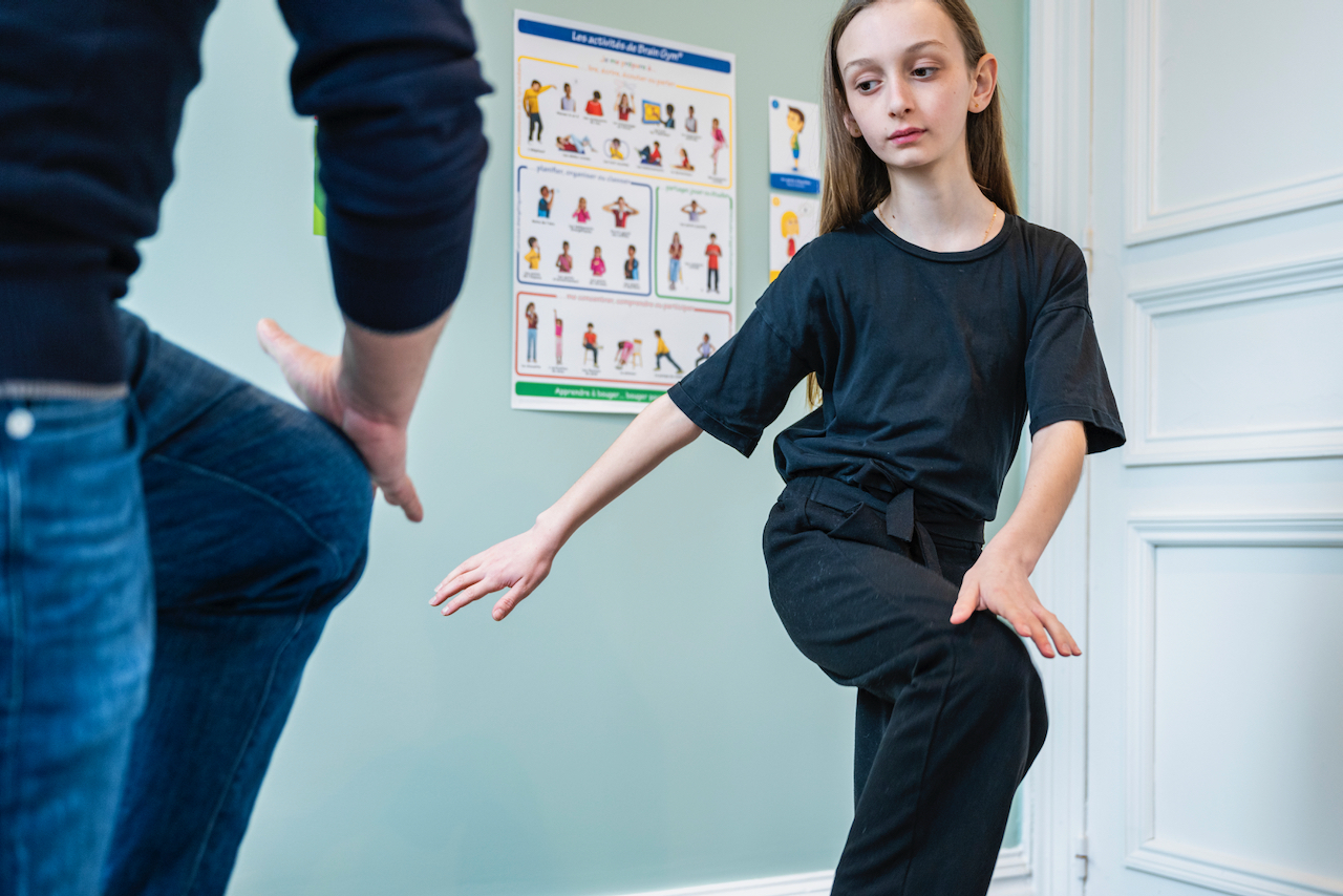 Une jeune fille debout dans une pièce. Elle est vêtue d'un t-shirt noir et d'un pantalon noir. Elle se tient sur une jambe, l'autre jambe étant pliée vers le haut, comme si elle effectuait un exercice d'équilibre. Une personne, partiellement visible à gauche de l'image, semble être en train de la guider ou de lui montrer quelque chose. Il y a également une affiche accrochée au mur à droite de la fille, illustrant différentes positions d'équilibrations.