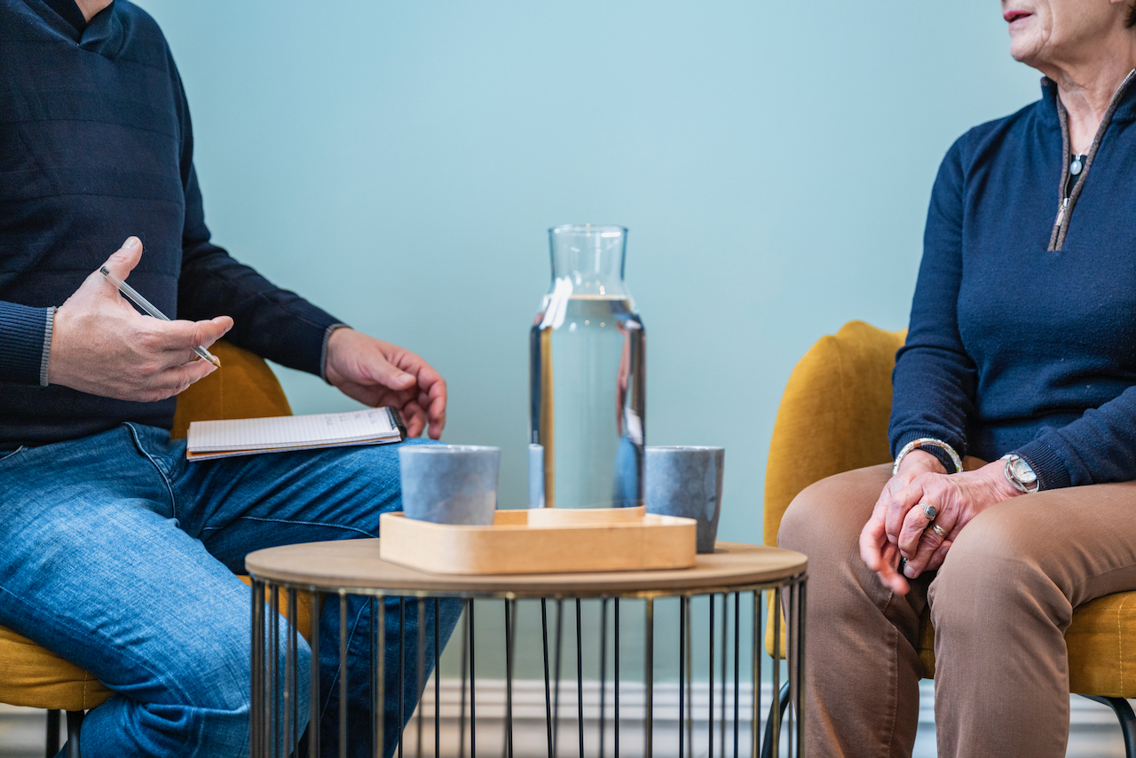 Deux personnes assises l'une en face de l'autre dans un cadre de consultation et de conversation. La personne à gauche est vêtue d'un pull noir et tient un carnet ou un cahier dans sa main gauche, tandis que la personne à droite est habillée en bleu foncé et semble écouter attentivement. Sur la table basse entre eux, il y a un pichet d'eau et deux verres, ce qui suggère que la réunion est informelle et professionnelle. Le décor est sobre avec un mur bleu clair en arrière-plan.